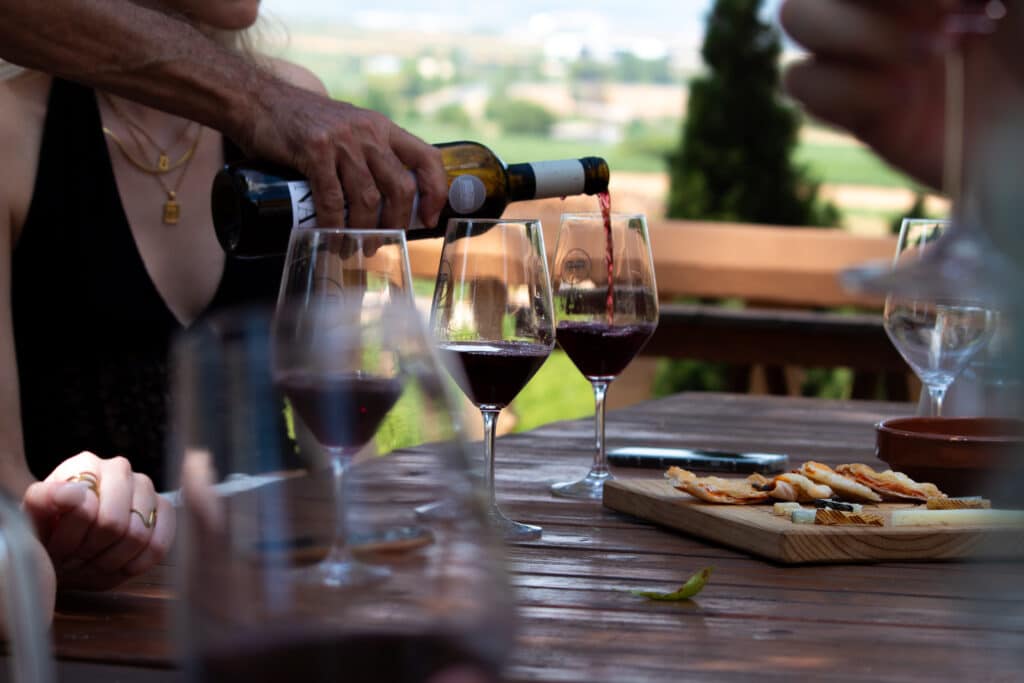 photo of wineglasses being filled with red wine on a table outside, there are two people sitting at the table